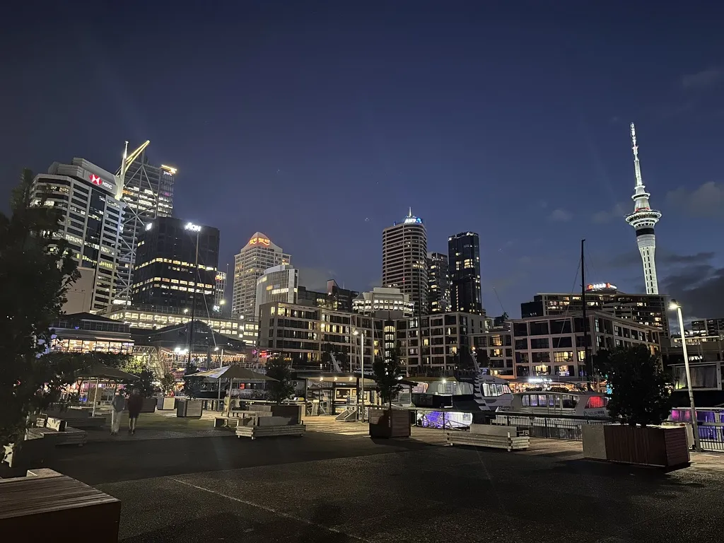 Auckland's waterfront at night, the lit Sky Tower and downtown towers rising above a quiet boardwalk plaza