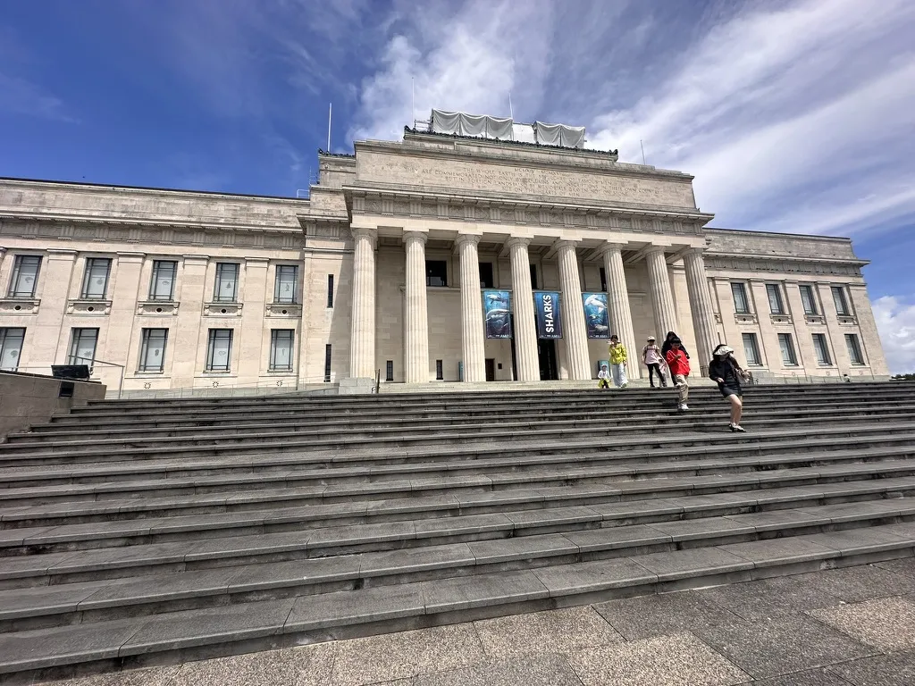 The Auckland War Memorial Museum, a grand neoclassical building with a columned portico, at the top of a wide flight of stone steps under a bright blue sky
