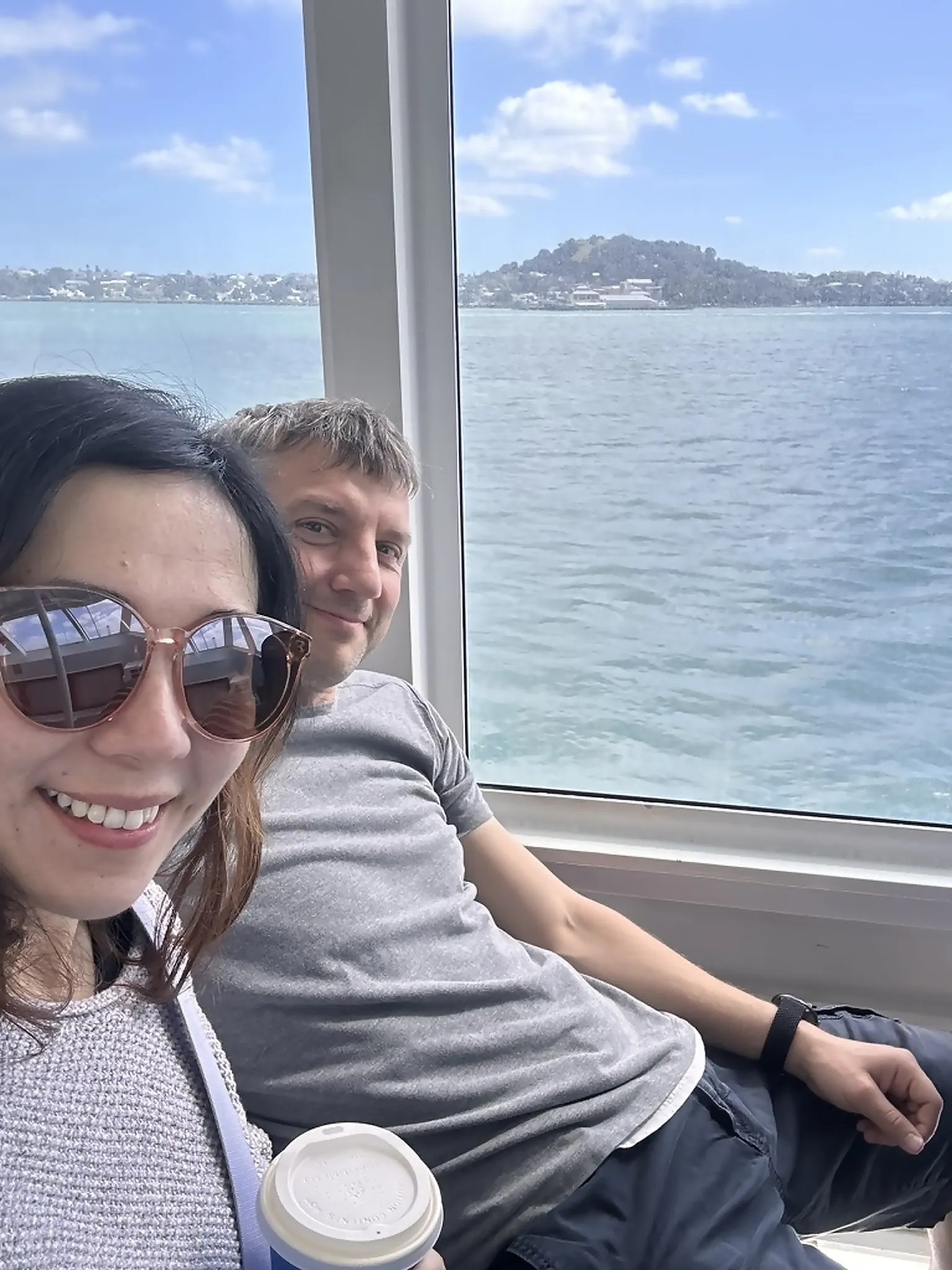 Kathryn in sunglasses holding a takeaway coffee smiles next to Karl on the ferry, the blue Hauraki Gulf and a green headland framed in the window behind them