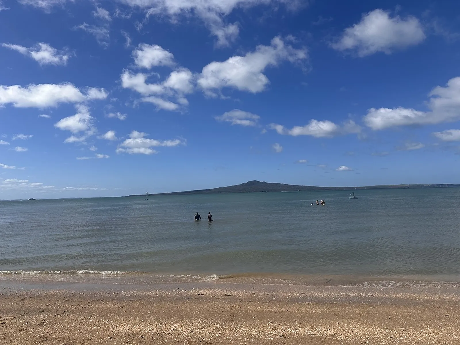 The low, wide cone of Rangitoto Island on the horizon seen from the sandy shore of Mission Bay, a few swimmers and a paddleboarder in the shallow blue water under a big cloud-dotted sky