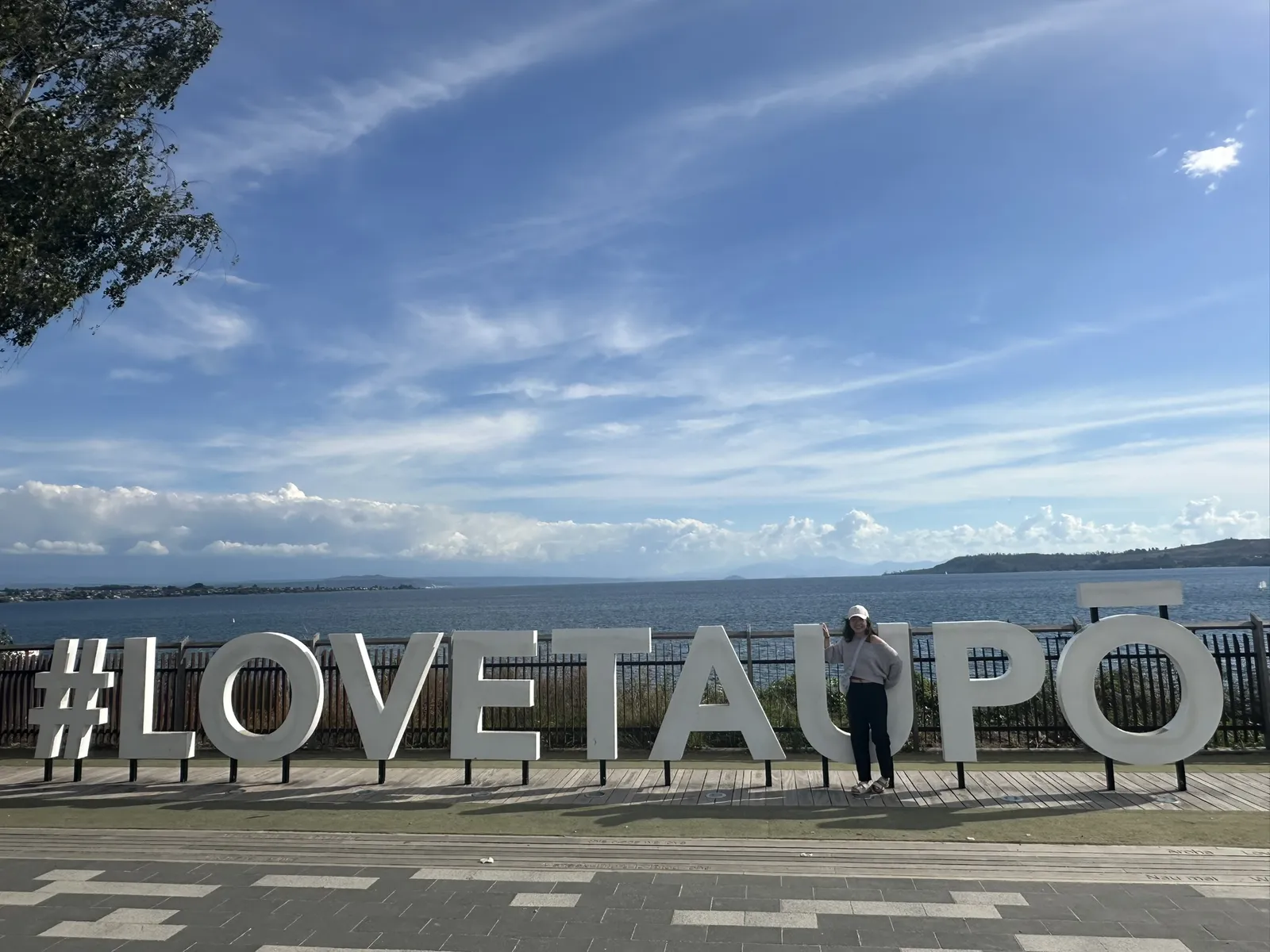 Kathryn in a white cap leaning on the giant white #LOVETAUPŌ letters on the lakefront promenade, the vast blue lake and distant ranges stretching out behind under a bright, wispy-clouded sky